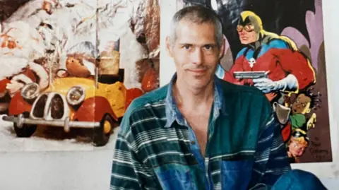 Supplied Nigel sitting in what appears to be a bedroom. He appears to be in his late 30s or early 40s. He is wearing an oversized green and blue shirt. There are children's television programme posters on the wall behind him.