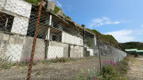 A view of the site, there is a metal fence in front of the remnants of a building on the cliff side.