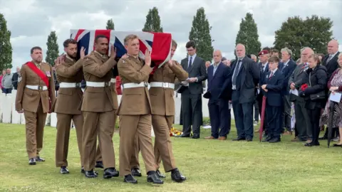 BBC/Olivia Richwald Military pallbearers in brown uniforms carry a coffin draped in a union flag. In the background there are mourners dress in smart clothing watching on.