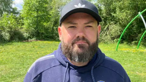 Mike Jones poses for the camera. He has a full dark beard and is wearing a black baseball hat and a blue Farnborough FC hoody.
