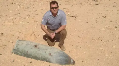 Ben Remfrey wearing a grey polo shirt and brown trousers kneeling down on sandy ground next to the shell of an explosive.