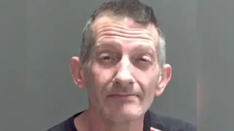 Nigel Halt is in the middle of the image looking towards the camera. He has short dark grey hair, and is wearing a black top. He is stood in front of a grey background. 