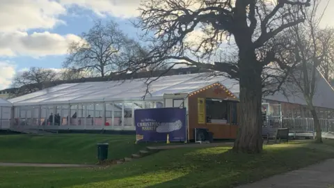 The ice rink in Imperial Gardens, Cheltenham. It is a large white marquee on a grassy field with festoon lighting and an advertisement sign for the Christmas Market. 
