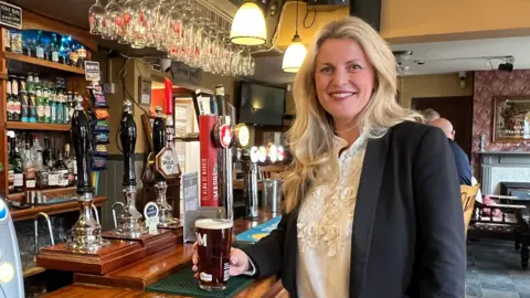 Emma is standing holding a pint at the bar. She has long blonde hair and is wearing a black jacket with an embellished cream blouse underneath.