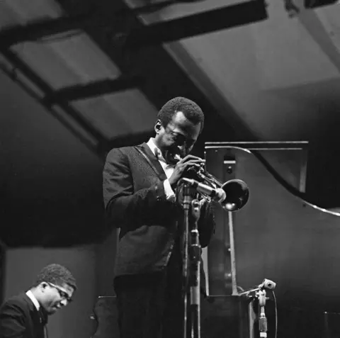 Getty Images Foto hitam dan putih dari Miles Davis memainkan trompet dengan Herbie Hancock di sudut memainkan keyboard.