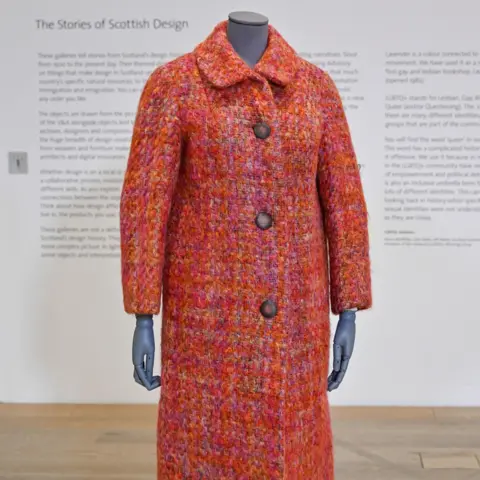 V&A Dundee A coat in fabric made by Bernat Klein - it is displayed on a Tailor's mannequin at the V&A. The 3/4 length coat is in a boucle tweed texture, woven in shades of orange, pink, red and purple with a rounded collar, bracelet-length sleeves and two big single-breasted buttons.  