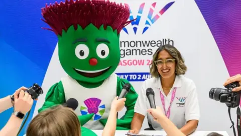 Clyde, an animated thistle mascot with green leaf-like skin and purple spiky hair sitting next to a smily lady wearing glasses and a white polo shirt. Behind them in a banner with the Glasgow 2026 logo on it. In front are people holding cameras and microphones.
