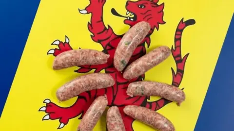 BRAMBLEBEE FARMS Eight uncooked sausages are arranged in a  circle on top of the flag. It features a red heraldic tiger facing left on a yellow stripe with a blue stripe on either side. 