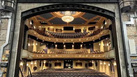 The image shows interior of the opera house from the stage looking out into the stalls. It is a large golden building with a number of seats. There is three layers to it.