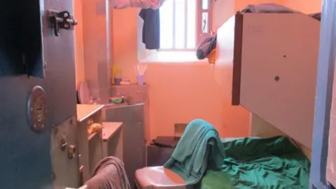 A cramped prison cell pictured from next to its metal door, showing bunk beds and chairs draped with towels and clothing.