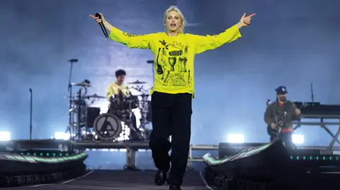 A woman wearing a yellow top and holding a microphone in her left hand is on stage, walking towards the camera. In the background behind her, both left and right, are band members playing.