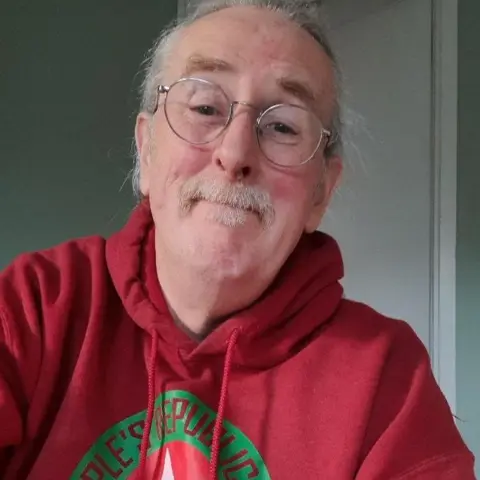 Paul Roberts Paul Roberts looking at the camera, half smiling and wearing a red hoodie. He is wearing glasses and his white/grey hair is tied back. He also has a moustache and small goatee.