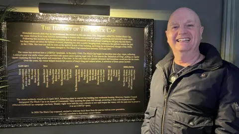 A bald man in a dark coat beams at the camera. He is standing next to a framed piece of writing entitled "The history of The Black Cap"
