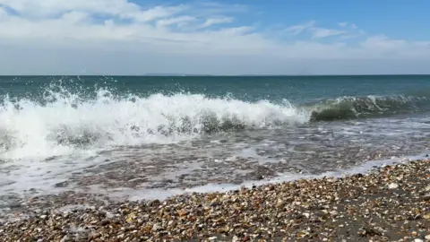 George Carden/BBC Waves gently crashing on bracklesham bay's pebbly beach. In the distance beyond the calm seas is the outline of the Isle of Wight