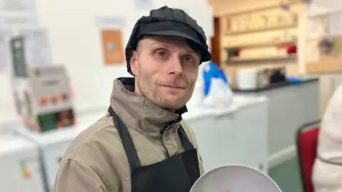 Darren Jones, a man wearing a beige coat and a black apron over the top of it as well as a black catering hat, is smiling and looking at the camera whilst holding a bowl which is tilted towards the photographer. He is indoors in what appears to be a room with a number of cupboards and worktops.