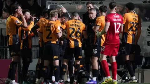 PA Media Cambridge United players, in black and amber shirts and a goalkeeper wearing red, stand in a huddle speaking to their manager. 