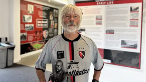 Adrian Pope in a white football shirt with a red and black crest and a large sponsor's logo. He is carrying a small metal bust of a man in a suit. An exhibition is visible behind him.