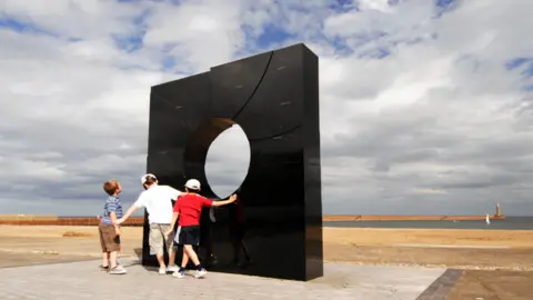 Andrew Small Three young boys stand next to a large, polished slab of black granite looking out towards a harbour with a lighthouse at the far end. 