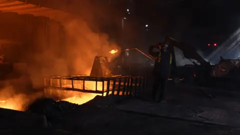 BBC A steelworker at Blast Furnace 4 tests the liquid iron that glows orange as it emerges from the furnace