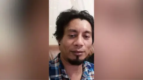 Ruben Alexandre Guimares who had short black hair and a black goatee. He is wearing a blue and orange gingham shirt and sitting on a brown sofa. 