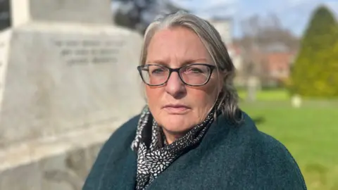 Hilary wears a blue coat and black and white scarf, with black rimmed glasses. She's looking to camera and standing in front of the white memorial to the Princess Alice victims.