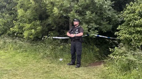 Policeman with police tape at the scene of the incident. Green bushes are around the cordon and a path is visible. 