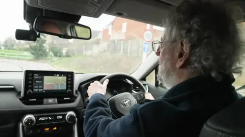 Photograph of Turner driving, taken over his shoulder. You can see the back of Turner's head - he has short, curly grey hair and a beard. He is wearing a pair of thin-framed glasses, and a dark blue fleece. The interior of his car is black, with a silver Toyota logo on the wheel. Outside the windscreen, you can see Turner is driving down a residential street. 