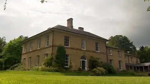 A large two -storey country house, with a garden in the front and the trees behind it.
