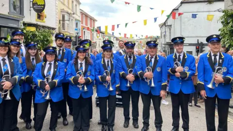 A band wearing blue blazers with black lapels and match ties, as well as matching black caps are seen in the street. Each member of the band pictured is carrying a silver trumpet.
