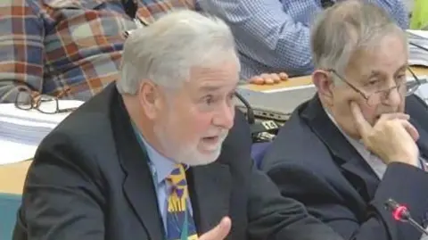 FoDDC Councillor John Francis, who has grey hair and a grey beard, sits in a council chamber wearing a dark suit and a yellow, blue and green patterned tie. He is turned slightly away from the camera, looking towards a microphone during the meeting.
