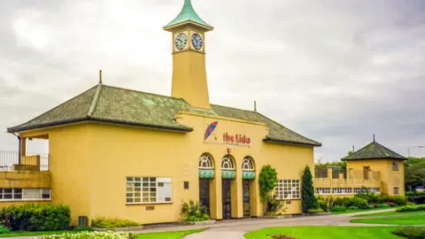 Chris Porsz Picture of the lido- a yellow-ish building with a green roof with grass in front of it, with The Lido written in red at the front of the building.