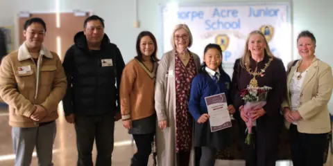 Charnwood Borough Council A row of people posing for a photo in a junior school hall alongside Felicity with a certificate, including smiling family members and the Mayor in her chain of office holding a bouquet of flowers