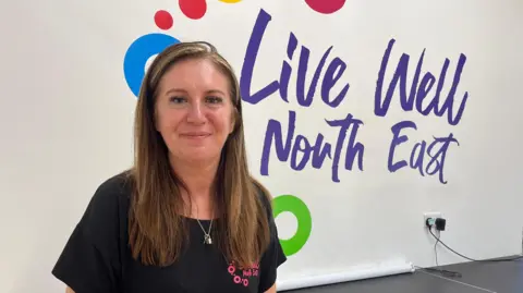 BBC/Andy Watson Debra Cumiskey wears a black top with a bright pink logo stands in front of a white wall emblazoned with the logo Live Well North East which is written with a playful font in a purple colour. 