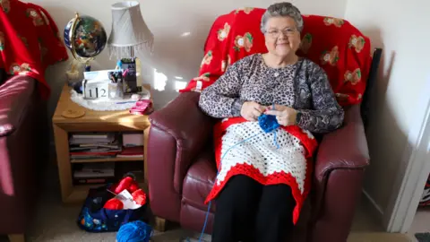Gail Foster A woman with short grey curly hair is sat in a red leather chair with a ball of wool and a white and red crochet blanket.