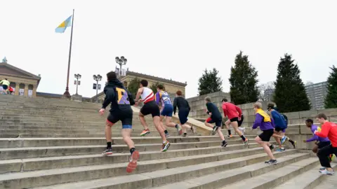 Getty Images Group of people running up the grey steps of the Philadelphia Museum of Art, many of them are in running kit. A flag flies from a pole to the left, with the pillars of a stone building visible on the left.