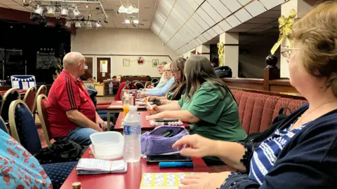 Six men and women dressed in casual clothes are sitting around rows of red-topped tables in a function room of a club. A stage can be seen in one corner with spotlights hanging from the ceiling.