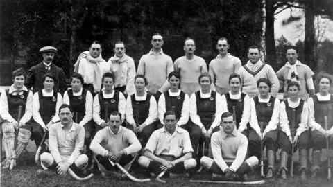 BPEOSA collection A black and white image of a large group of people. A row of men are sitting down, at the front, with their legs crossed, then a row of women in gym slips, sitting down, and then a row of men behind them all standing. 