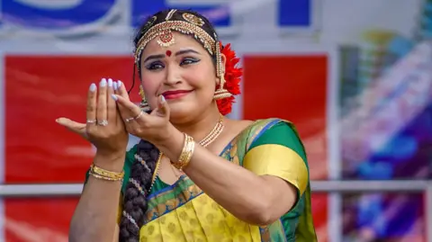 A woman with her hair in a long plait, wearing a green and yellow sari and a gold beaded headpiece, holding her arms outstretched in a dance