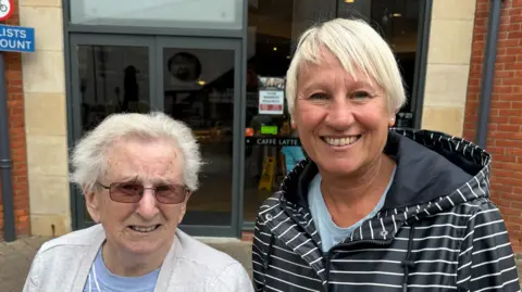 Sharon and her mother Valerie pose for a picture outside the front of a cafe in Didcot. On the left, Valerie, aged around 70-90, is wearing a light blue t shirt with an open white cardigan over the top. She has white hair and sunglasses. Her daughter Sharon has a light blue t shirt and a large blue and white striped raincoat with a hood. She has short, straight white hair.