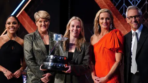 PA Media Beth Mead poses with The BBC Sports Personality of the Year Award alongside presenters Alex Scott, Clare Balding, Gabby Logan and Gary Lineker during the BBC Sports Personality of the Year Awards 2022. Mead, in the middle, has long blonde hair and is wearing a grey blazer. She is holding the large silver trophy, which is in the shape of a film camera, at waist-height.
