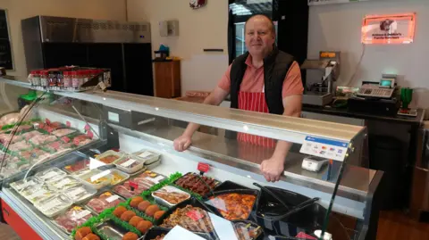 Butcher Andrew Garner is wearing  a red apron and is standing behind his counter full of meat.