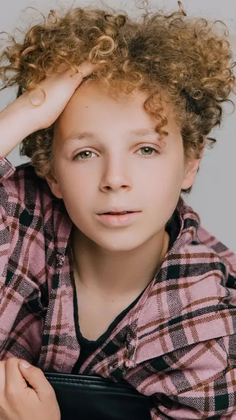 FayAndrea Photography A professional-style headshot of a young boy looking into the camera while pushing a mop of curly hair away from his forehead.