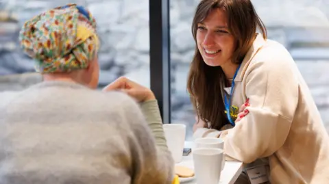 UK for UNHCR Two women sitting at a table at a pop-up cafe chatting over cups of tea and biscuits