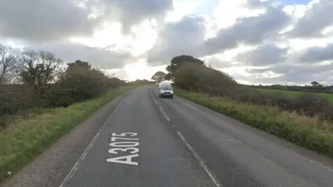 The A3075 near Rejerrah in Cornwall on a sunny but cloudy day. Fields, hedges, trees and grass verges are on both sides of the road. A silver van and silver car are driving on the road.