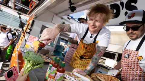 PA Media Ed Sheeran signs a head of broccoli in hot sauce outside a Sainsbury's supermarket in London Colney to promote his Tingly Ted's hot sauce brand