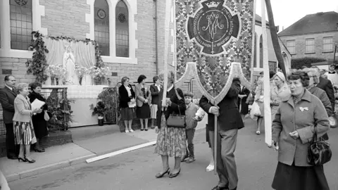 Tim Smith A black and white photograph of people marching past a church with one man's head and shoulders obscured by a religious banner he is holding.