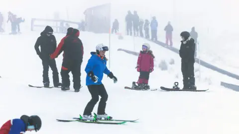 Skiers and snowboarders of various ages practise on a snowy slope in foggy conditions, with instructors and groups scattered across the background.