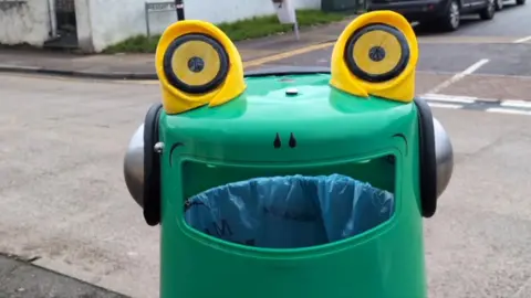 A close-up of a green bin with two yellow eyes on the top and a set of headphones. The opening of the bin is designed to look like a mouth, with a nose drawn above it.