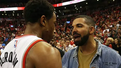 Pictured in 2016,  Drake congratulates DeMar DeRozan, when they were on happier terms. Drake, on the right, is wearing a brown tshirt and blue denim jacket, and is speaking to DeMar, who is wearing a white Toronto Raptors jersey. The background has fans in the arena in Toronto.