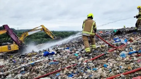 Staffordshire Fire and Rescue Service Two firefighters holding fire hoses spraying water at a fire on an area of rubbish at a landfill site.